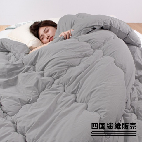 一位女子舒適地躺在灰色雙人厚暖冬被中，被子表面呈現柔軟蓬鬆的質感，邊緣有明顯的縫線，被子填充物為中棉1.2kg。