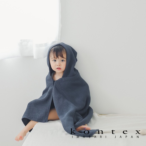 一名穿著深藍色針織純棉連帽浴巾的嬰兒，坐在白色背景前的床墊上。