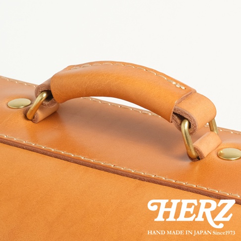 HERZ 手作皮革橫式書包提把特寫，皮革材質，配有金色金屬扣環，下方可見品牌標誌。