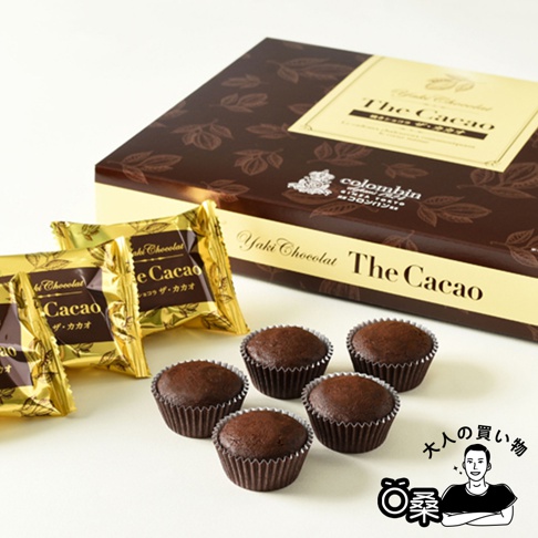 一個印有金色「The Cacao」字樣和葉子圖案的深棕色禮盒，旁邊擺放著幾個獨立包裝的金色巧克力菓子，以及五個裝在銀色紙杯中的深棕色巧克力馬芬。