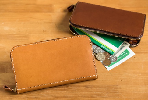 兩個職人手作皮革長夾，一個為淺棕色，另一個為深棕色，皆為拉鍊式設計，表面有細緻的縫線。淺棕色長夾前方，散落著錢幣、紙鈔和綠色存摺。