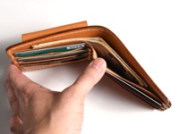 手持一棕色皮革雙折短夾，夾內有鈔票、信用卡及零錢袋，皮革邊緣縫線清晰可見。