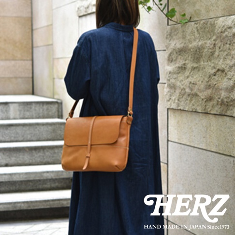 日本HERZ職人手作皮革細扣帶馬鞍包，焦糖色皮革，模特兒身穿牛仔藍長洋裝，側身斜背展示包款。