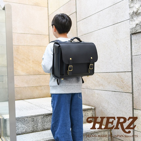 男孩背著黑色皮革橫式書包，書包上有金屬扣環，下方有HERZ品牌字樣。