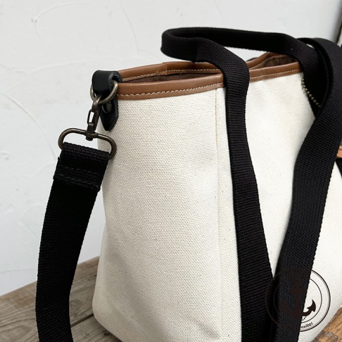 高島帆布多口袋手提托特包，以米白色帆布製成，配有棕色皮革飾邊與黑色提把和可拆卸斜背帶。包包上有金屬扣環和 D 型環，展現簡潔實用的設計。
