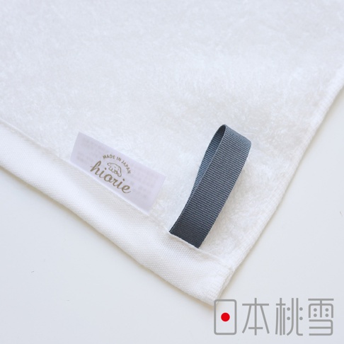 一條純白色、觸感柔軟的迷你毛巾，帶有深灰色織帶掛耳，織帶末端縫有「hiorie」品牌標籤，標籤上印有「MADE IN JAPAN」字樣。