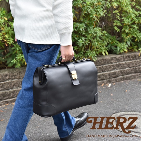 黑色皮革製成的職人手作公事包，配有金色鎖扣和提把，由一位身穿牛仔褲和白色毛衣的人士手提。