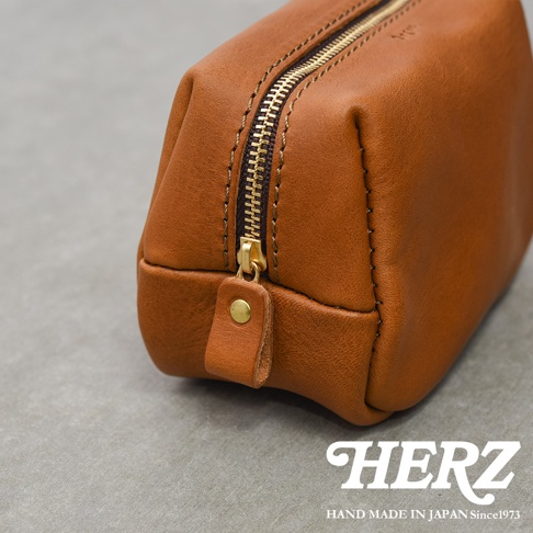 一個棕色皮革手工收納包的近距離特寫，鏡頭聚焦在黃銅拉鍊與皮革接縫處，收納包表面具有細膩的天然皮革紋理，底部右下角印有日系手工品牌HERZ的白色標誌與創立年份字樣，整體呈現簡約質感與職人手作工藝。