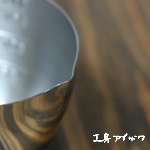 不鏽鋼長柄量杯，銀色，帶有刻度，杯緣有導流設計，放置於木紋背景上。
