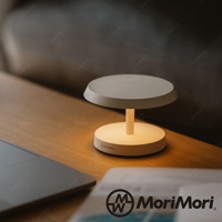 米白色圓盤燈座配上圓盤燈罩，中間由一根短柱支撐，燈具散發柔和暖黃光，放置於木紋桌面，旁邊有筆記型電腦。