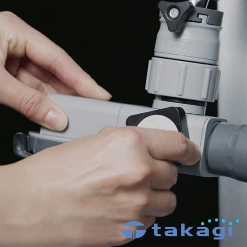 雙手正在操作灰色塑膠材質的水龍頭分支蓮蓬頭，連接處有黑色按鈕和白色標籤，背景為黑色。