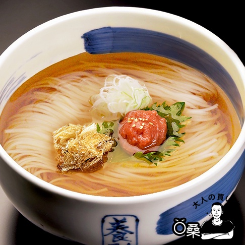 碗中盛有清澈的湯頭，浸泡著細緻白色的烏龍麵。麵上擺放著一團紅色帶有芝麻的醬料，一撮金黃的炸物，以及切細的蔥花和綠色葉片。碗邊緣有藍色筆觸的裝飾。