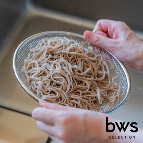 一雙手正拿著一個不鏽鋼瀝水籃，裡面裝滿了煮熟的蕎麥麵，瀝水籃放在流理台上方，下方可見部分廚房的細節，右下角有「bws SELECTION」的品牌標誌。