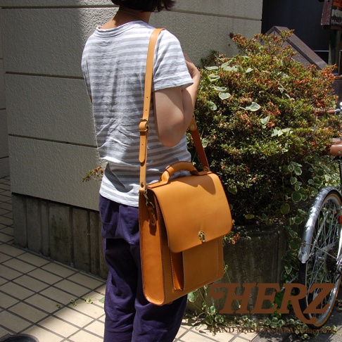 一個身穿條紋上衣和深藍色褲子的人，斜挎著一個棕褐色的皮革後背包。背包設計簡潔，帶有提把和金屬鎖扣，材質看起來是真皮。