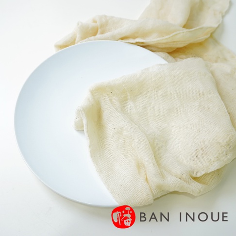 一張白色盤子上疊著一塊米白色、網狀織紋的有機蚊帳棉廚房拭巾，拭巾的邊緣自然垂落。