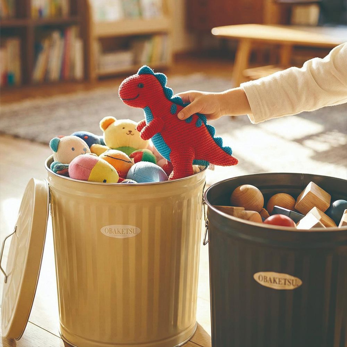 一個孩子的手正從一個駝色的 OBAKETSU 垃圾桶裡拿出一隻紅藍色編織的恐龍玩具，桶裡還裝著其他絨毛玩具，旁邊還有一個深色的 OBAKETSU 垃圾桶裝著木製玩具。