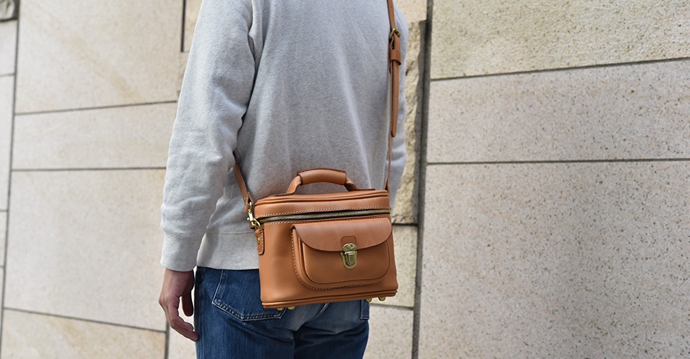 焦糖色皮革手提斜背包，側面有口袋和金色鎖扣，頂部有拉鍊和提把，可肩背。