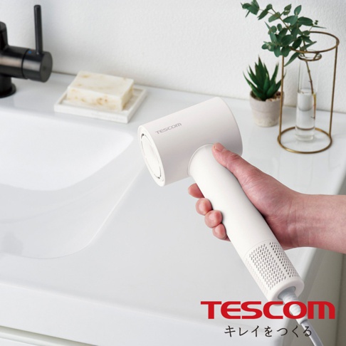一只手拿著一個白色的 TESCOM 負離子吹風機，吹風機上有電器品牌字樣，吹風機的後方有網狀的設計，背景是一個浴室的洗手台，台上還有香皂和植物裝飾。