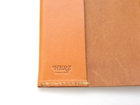 職人手作皮革附繩書套，淺棕色天然皮革，帶有壓印的「HERZ」品牌標誌及「MADE IN JAPAN」字樣，邊緣縫線工整。