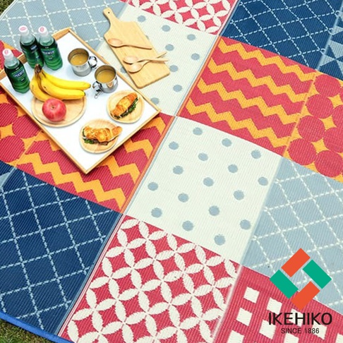 防水 PP 材質的野餐墊，由多種幾何圖案拼接而成，包括紅白色花紋、黃紅色鋸齒紋、藍色菱形格紋、白色圓點紋及紅色圓點紋。墊上放有裝有香蕉、蘋果、麵包和飲品的餐盤，以及木質砧板和木勺。