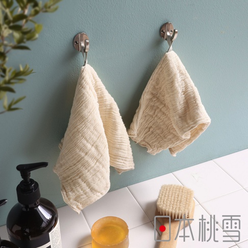 日本桃雪的立體褶皺柔綿紗布洗臉巾，掛在牆壁的掛鉤上，左邊是一瓶黑色瓶身的洗手乳，右邊是洗臉巾旁放著一顆琥珀色的肥皂和一把木柄刷毛的洗臉刷。