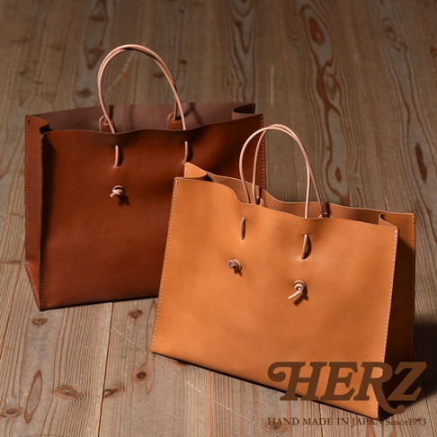 兩款職人手作皮革提袋，一款為深棕色，另一款為淺棕色。提袋均為方形設計，使用皮革材質，配有兩條細長的皮革提把，並以細繩打結固定於包身前方。
