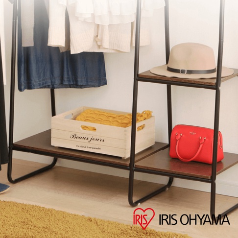 黑色金屬框架搭配木紋層板的吊掛衣架，展示有藍色洋裝、針織衫、木質收納盒（內含黃色毛毯）及紅色手提包，上方層板放置一頂米色寬邊帽。