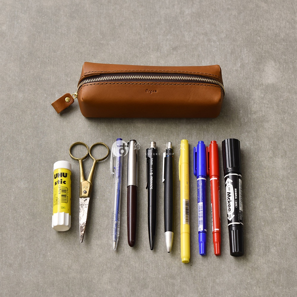 職人手作皮革Organ箱型收納筆袋，咖啡棕色，附拉鍊和皮革拉環，下方擺放著一支黃色UHU膠水、一把金色小剪刀，以及多支不同顏色筆。