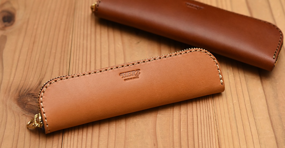 兩個棕色皮革筆袋，上面壓印著HERZ品牌標誌，放置在木紋桌面上。