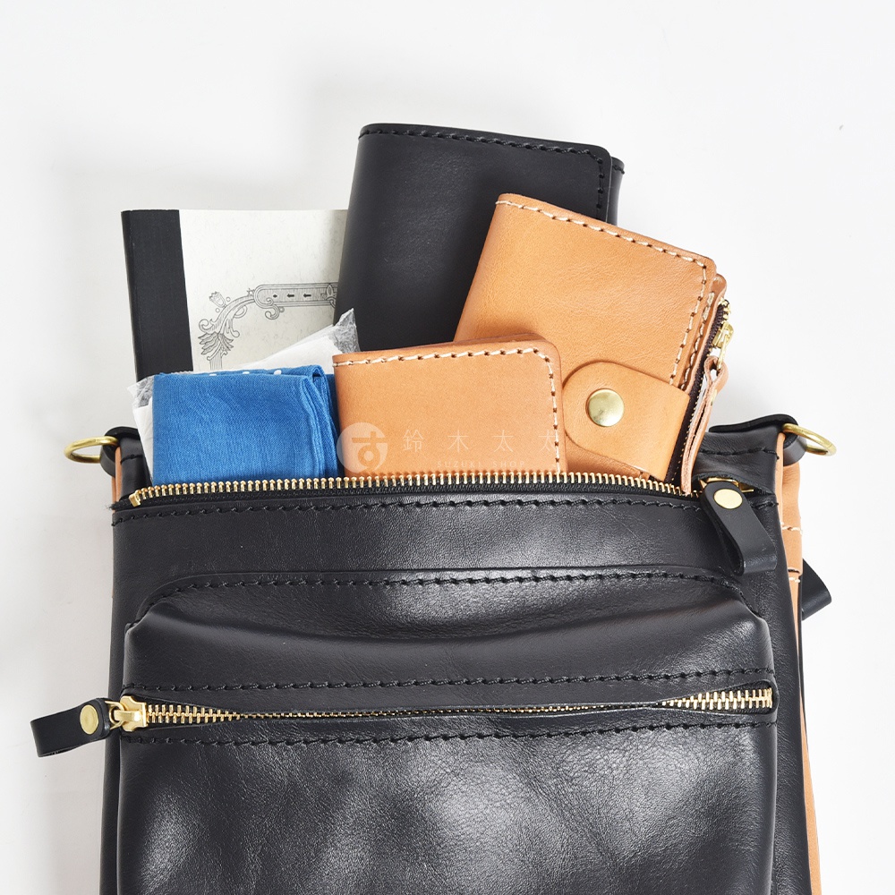 黑色皮革斜背包，內部裝有筆記本、藍色布料、棕褐色皮革錢包和卡夾，搭配金色拉鍊和金屬扣。