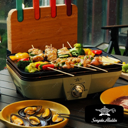 淺綠色迷你烤盤上烤著烤肉串，有雞肉、青椒、紅椒、櫛瓜和花椰菜，旁邊有木製竹籤架，架上插著彩色竹籤。右下角為黃色盤子，盤中盛有淡菜。