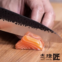 一把黑色錘紋刀刃的主廚刀，正在切割木板上的三片鮭魚，刀刃呈現大馬士革紋路。