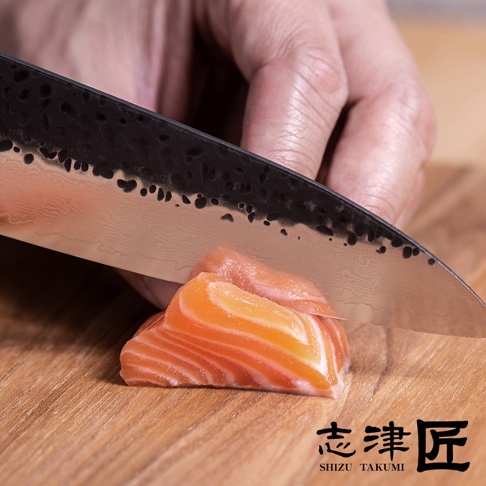 一把黑色錘紋刀刃的主廚刀，正在切割木板上的三片鮭魚，刀刃呈現大馬士革紋路。