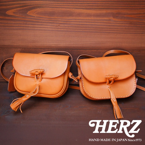 兩款職人手作皮革流蘇側背包，以天然皮革製作，呈現溫潤的焦糖色澤。包包設計簡約，翻蓋處以皮革繩結固定，下方裝飾著皮革流蘇，展現細膩的工藝。