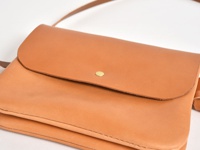 職人手作皮革直式隨行袋，採用光滑的淺棕色皮革製成，帶有翻蓋設計和一個黃銅色金屬鈕扣。