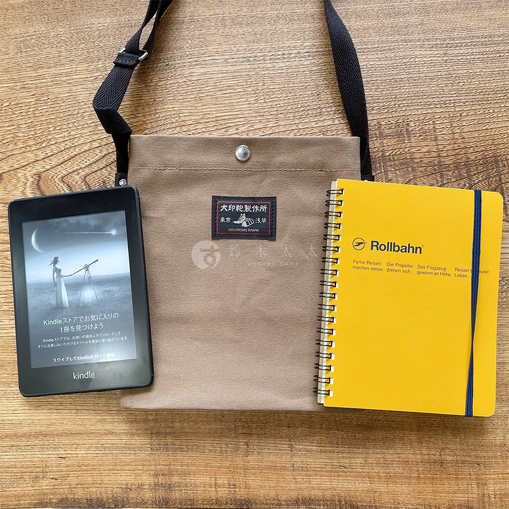 一個米色帆布平板收納側背包，左側放著一台黑色 Kindle 電子閱讀器，右側擺著一本黃色 Rollbahn 筆記本，背景為木紋桌面。