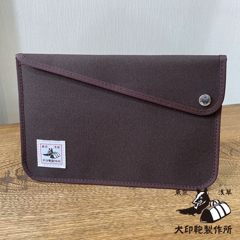 深棕色純棉帆布平板收納包，配有斜織帶裝飾和按鈕開合。左側有一個印有日文圖標的白色標籤。