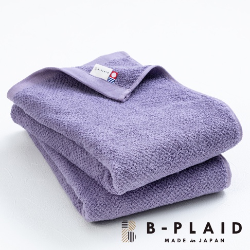 疊放的紫色薄手鱗紋浴巾，表面有細緻的織紋，其中一條浴巾的角落處可見白色品牌標籤，標籤上有「B-PLAID」字樣及公司Logo。