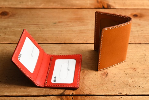 職人手作皮革雙折票券收納夾 (KC-32)，展示一個打開的紅色皮革收納夾，內有透明夾層，以及一個站立的棕色皮革收納夾，邊緣有白色縫線，兩者皆置於木紋背景上。
