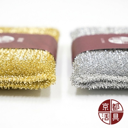 這是一張近距離拍攝的商品特寫照，畫面中並排放置兩塊不沾鍋專用菜瓜布，左側為金色款式，右側為銀色款式，兩者皆帶有細緻的金屬光澤紋理，中間分別綑綁著深紅色的品牌包裝紙條，右下角印有紅色的京都活具圓形標誌，整體呈現簡潔明亮的廚房清潔用品質感。