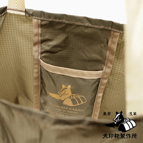 犬印鞄環保折疊收納購物袋，米色織帶提把，內部有深褐色網格內袋，袋上印有金色狐狸與行李箱圖案，並標示「INUIRUSHI KABAN made in Japan Pocket Tote」。