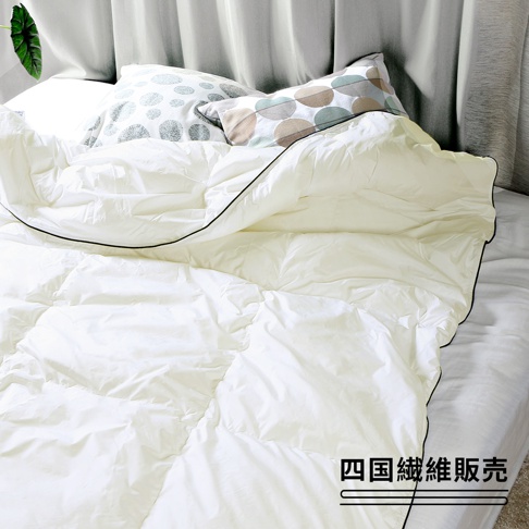 一張鋪在床上的白色雲絨被，被子表面有方形格紋縫線，四周鑲有黑色滾邊。背景是灰色的床頭板和兩顆枕頭，其中一顆枕頭印有圓形圖案，另一顆枕頭則有大小不一的圓形圖案。