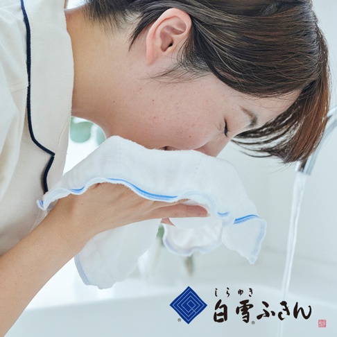 白雪肌洗臉巾，女子正用藍色滾邊的白色毛巾洗臉，臉部皮膚有雀斑，背景為浴室水槽和龍頭。