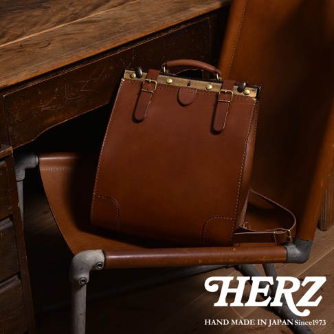 棕色皮革復古口金後背包，具備頂部提把、可調節式肩帶、金屬扣環及四個角落的加固設計，擺放在椅子上，旁邊是木製桌子。