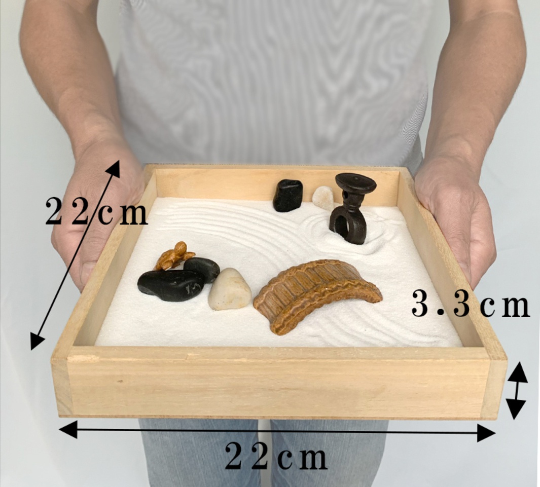 日式枯山水模型，木質淺盤盛裝白色細砂，砂上佈有耙痕。盤內有黑色、白色鵝卵石，一座棕色拱橋模型，及一座黑色榫卯結構小模型。
