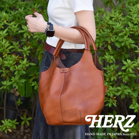 這是一個棕色皮革製成的圓形肩掛托特包，包身有明顯的縫線裝飾，提把為皮革材質。