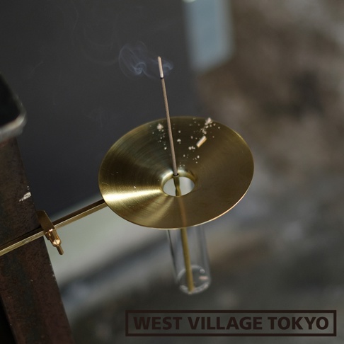 黃銅製的圓形線香架，帶有可夾持的設計，中間有玻璃管，上方插著一炷燃燒的線香，散發出陣陣煙霧。