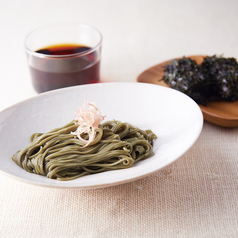 一碗綠色的蕎麥麵，上面點綴著粉紅色的薑絲，旁邊有一個玻璃杯裝著深色醬汁，另一邊有一個木盤裝著兩團海苔。