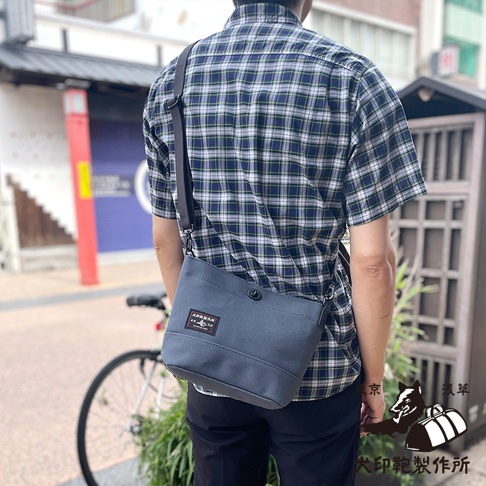 一位男士身著藍綠色格紋襯衫，背著一個灰藍色純棉帆布迷你側背包，包上有「犬印製作所」的標籤，背景是日本淺草的街道景色。