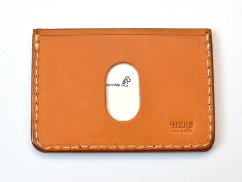 職人手作皮革橫式票券收納夾，採用棕褐色皮革製成，正面中央有橢圓形透明視窗，右下方壓印有「HERZ MADE IN JAPAN」字樣，邊緣以白色縫線手工縫製。